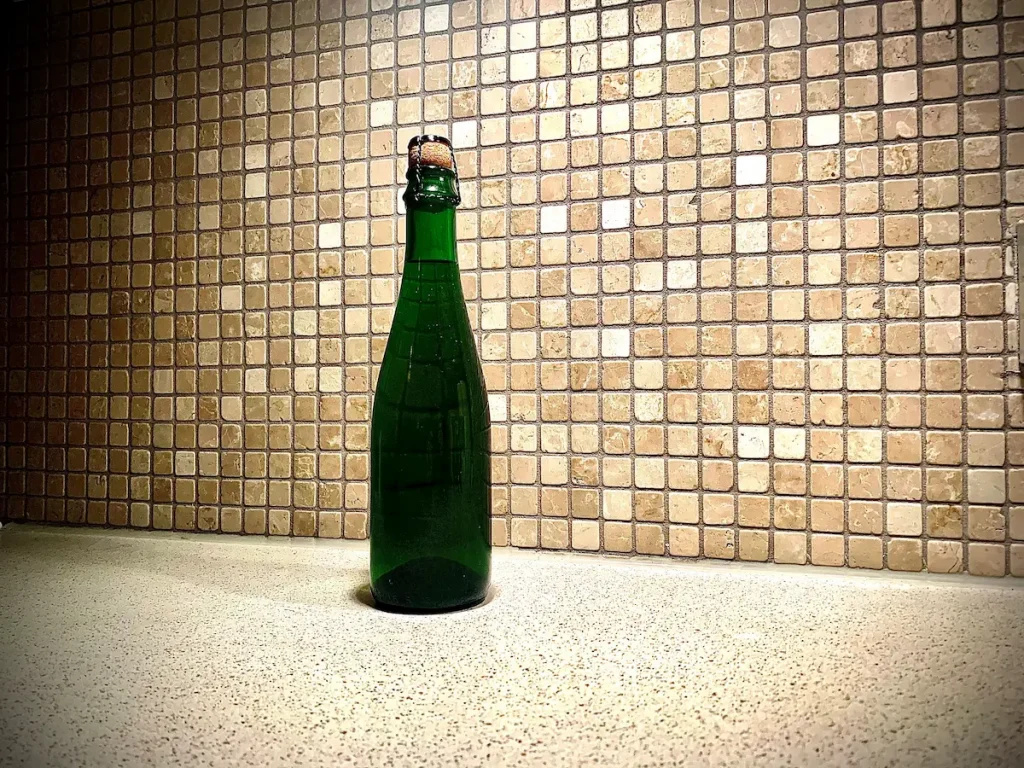 En grønn flaske med kork og metalltråd står på en lys kjøkkenbenk foran en vegg med små beige mosaikkfliser. Flasken er typisk for belgisk surøl som geuze eller lambic.